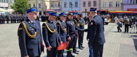 Moment dekorowania strażaków stojących w szeregu przez innych strażaków. W tle zbiórka strażaków, czerwone samochody i zabytkowe kamienice.