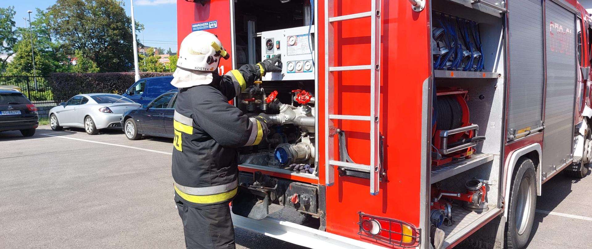Strażak w hełmie i ubraniu specjalnym obsługuje autopompę samochodu pożarniczego.