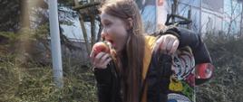 na fotografii dziewczyna siedzi na murku, trzyma deskorolkę i zjada jabłko

