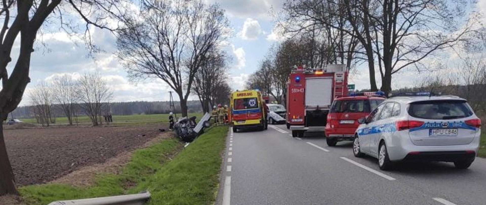 Po lewej widać pole, następnie samochód w rowie, na drodze są pojazdy straży pożarnej, karetka pogotowia i policja, droga zablokowana