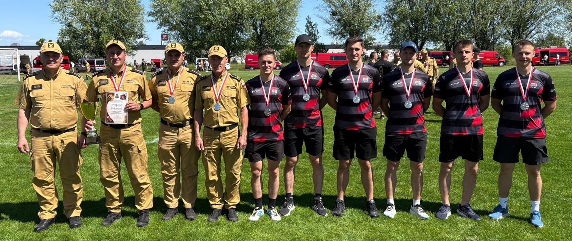 10 strażaków pozuje na boisku piłkarskim do zdjęcia, zaraz po uroczystości ogłoszenia wyników, strażacy zawodnicy z medalami oraz dyplomem, pośród nich komendant wojewódzki psp w łodzi oraz komendant powiatowy psp w Opocznie. W oddali widać wozy strażackie.