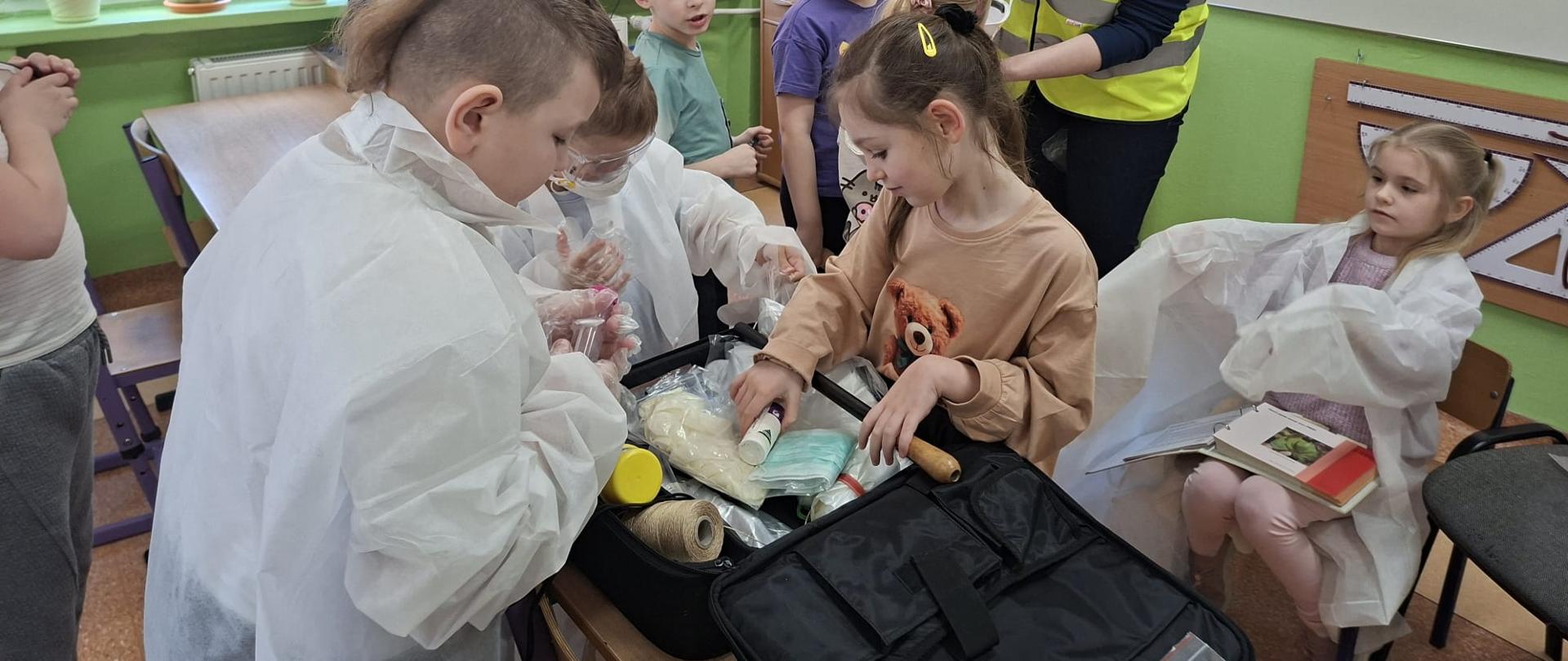 Zdjęcie przedstawia inspektora WIORiN prowadzącego spotkanie edukacyjnej w sali lekcyjnej pierwszoklasistów. Dzieci przymierzają ubrania robocze inspektorów WIORiN oraz przyglądają się zawartości torby inspektora WIORiN wypełnionej rożnymi środkami ochrony oraz narzędziami. 