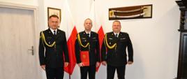 Zastępca pomorskiego komendanta wojewódzkiego Państwowej Straży Pożarnej stoi z trzema strażakami. Funkcjonariusz w środku trzyma czerwoną teczkę. 