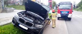 Czerne auto stojące na poboczu drogi. Uszkodzone auto z otwartą maską, w tle samochód bojowy Straży pożarnej.