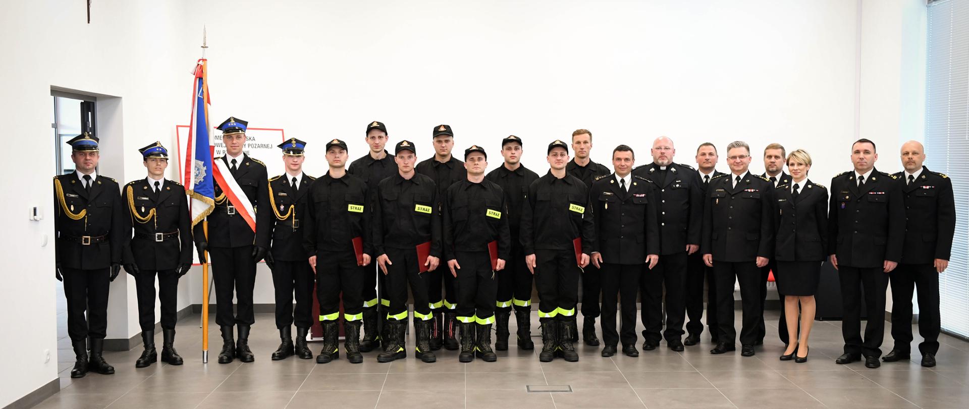 Zdjęcie przedstawia nowo przyjętych strażaków z reprezentacją Komendy Miejskiej Państwowej Straży Pożarnej w Rzeszowie, komendantem miejskim Państwowej Straży Pożarnej w Rzeszowie, zastępcą komendanta miejskiego Państwowej Straży Pożarnej w Rzeszowie, kapelanem wojewódzkim strażaków oraz pocztem sztandarowym na świetlicy Jednostki Ratowniczo-Gaśniczej Nr 3 w Rzeszowie. Wszystkie osoby przyjęły w postawę zasadniczą. 