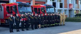 Strażacy stojący w szeregu na placu przed budynkiem komendy. Jeden ze strażaków trzyma na rękach flagę.