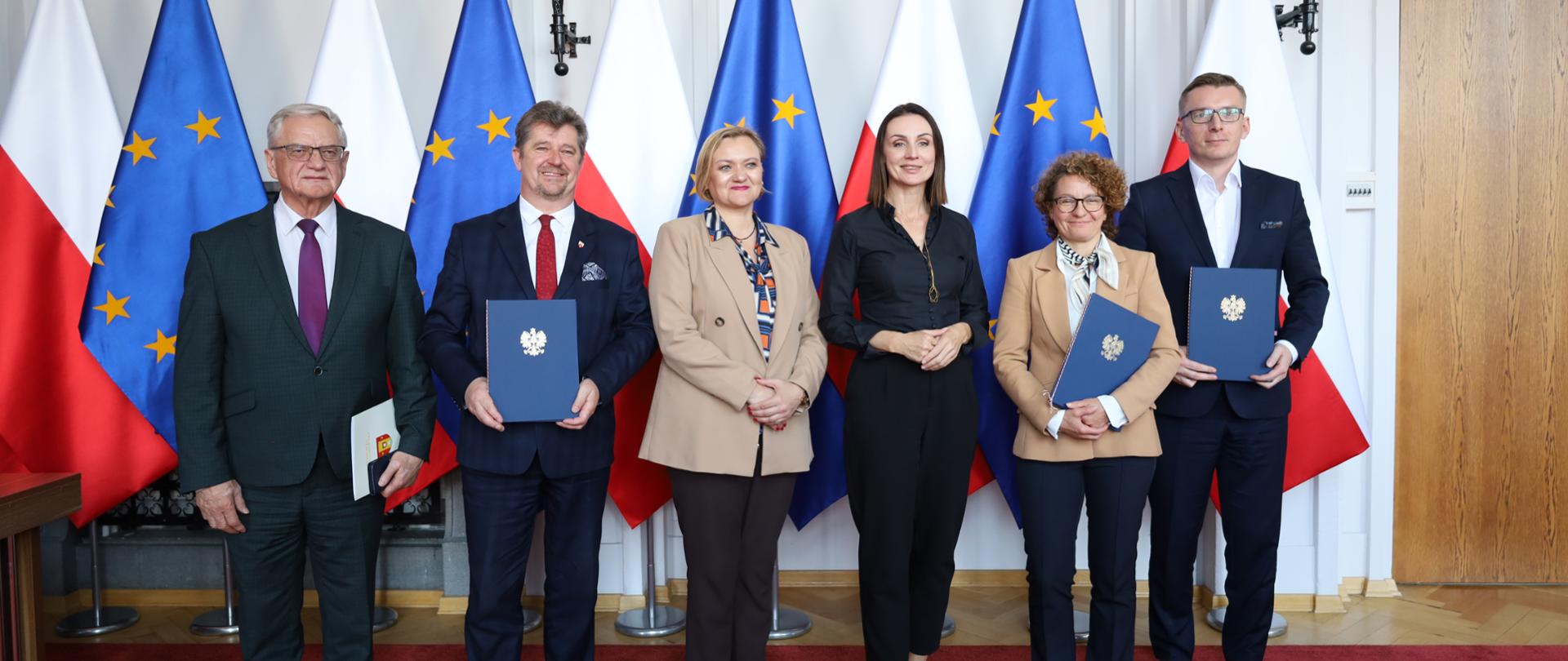 kilka osób trzyma w ręce teczki (podpisane umowy) i pozuje do zdjęcia na tle biało czerwonych i unijnych flag