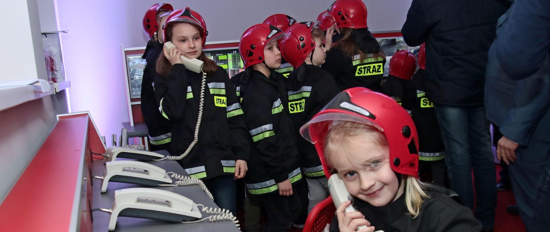 Zdjęcie przedstawia dzieci ubrane w hełmy i kurtki bojowe strażackie zgromadzone w pomieszczeniu z biurkiem, na który stoją telefony stacjonarne. Dwie dziewczynki prowadzą rozmowy trzymając słuchawkę przy uchu.