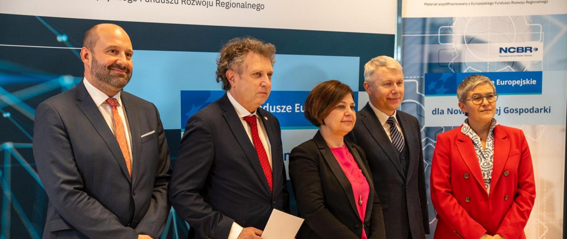 Przed ścianką w niebieskie wzory stoi pięć osób, z prawej wiceminister Mrówczyńska w czerwonym garniturze, obok niej kobieta w czarnej marynarce i trzech mężczyzn w garniturach.