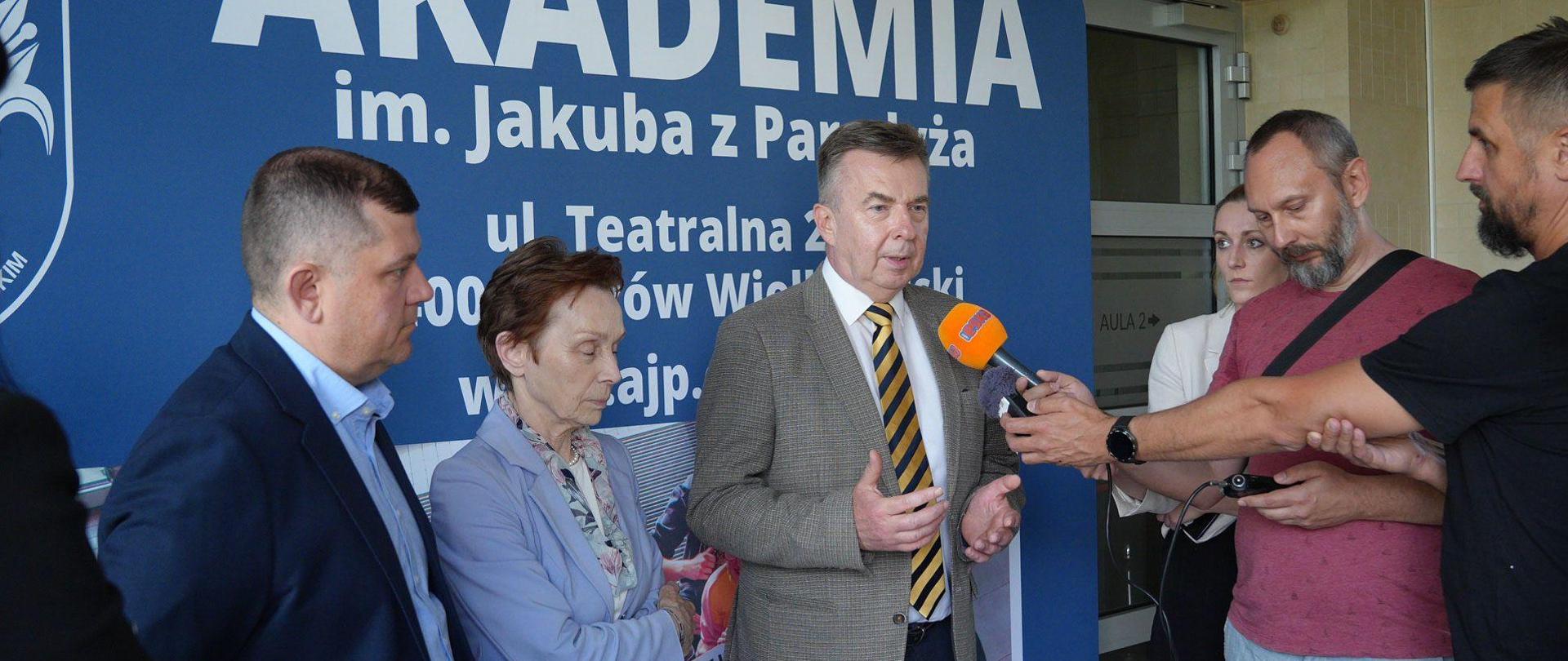 Pod niebieską ścianką z napisem Akademia im. Jakuba z Paradyża stoi minister Wieczorek, kobieta w niebieskiej marynarce i mężczyzna w garniturze, minister mówi do mikrofonu trzymanego w reku przez dziennikarza.