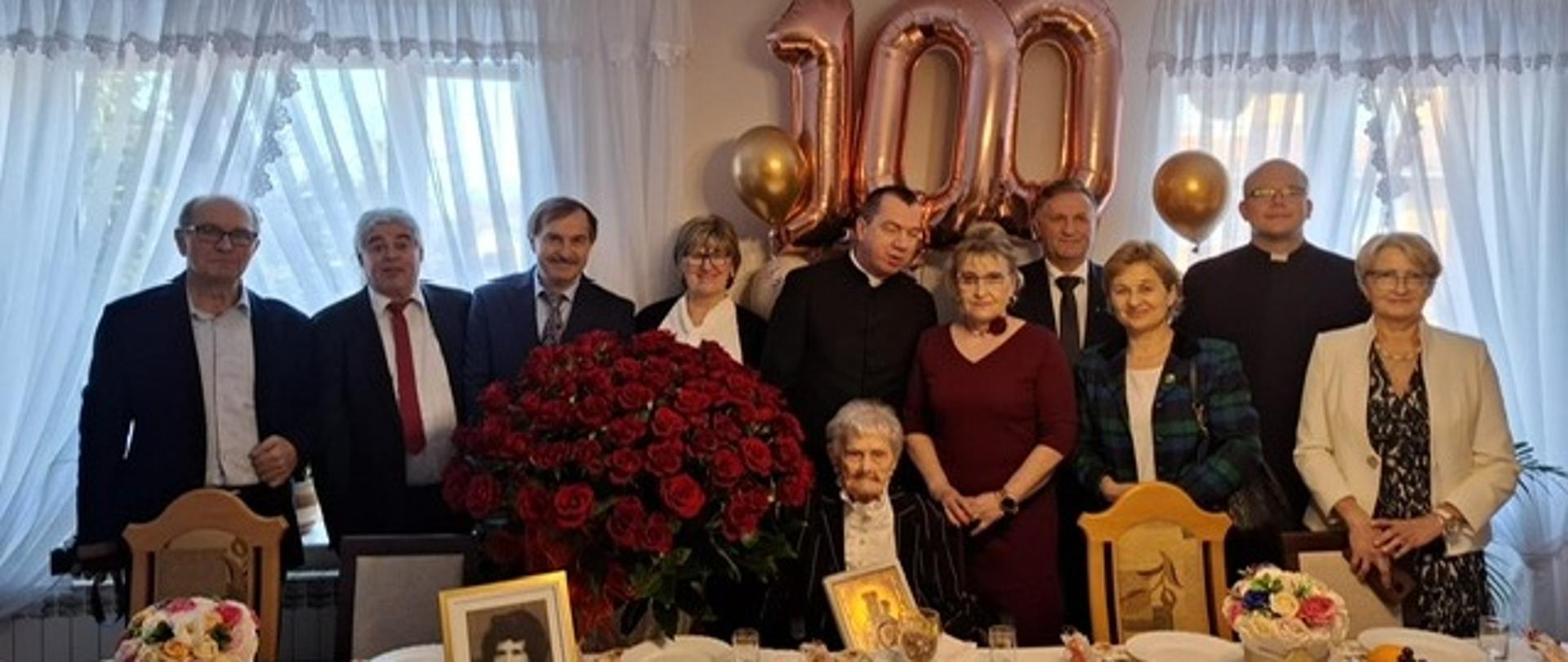 Jubilatka siedzi przy stole w otoczeniu dziesięciu uśmiechniętych gości – kobiet i mężczyzn w eleganckich strojach. Nad grupą wiszą duże, różowo złote balony tworzące liczbę 100. Na stole nakrytym białym obrusem stoi ogromny kosz czerwonych róż, mniejsze kompozycje kwiatowe oraz oprawione zdjęcie. W tle widoczne są jasne firanki oraz złote balony dekoracyjne.