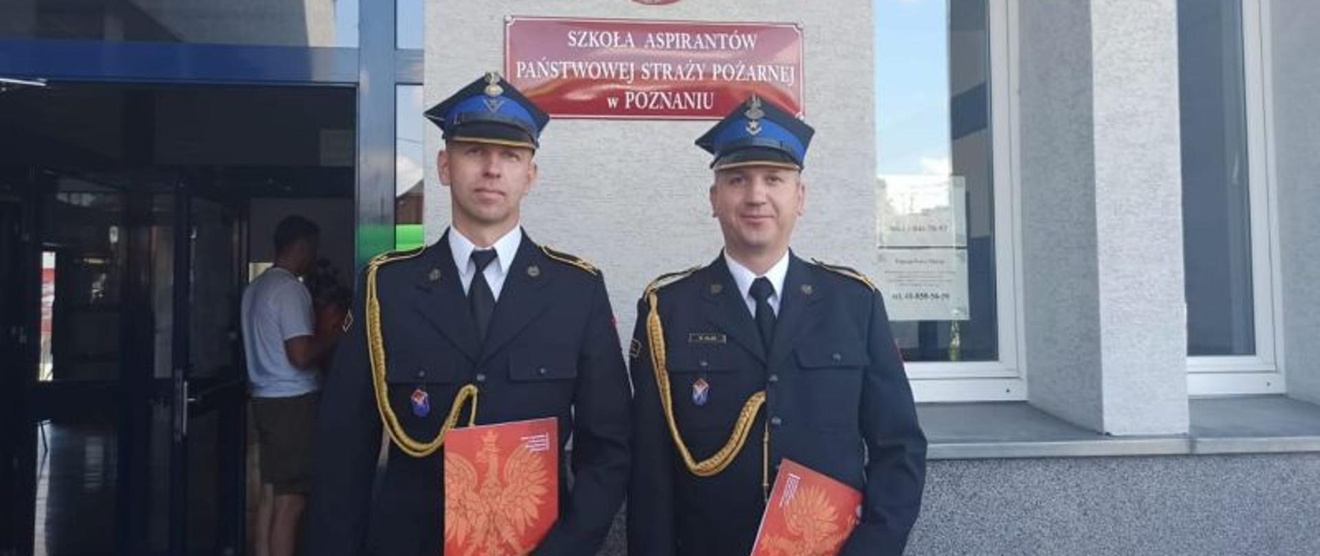 W centralnym punkcie zdjęcia stoi dwóch strażaków w galowych mundurach.Są to dwaj awansowani funkcjonariusze KP PSP Białogard. W tle widać czerwoną tabliczkę z nazwą szkoły.