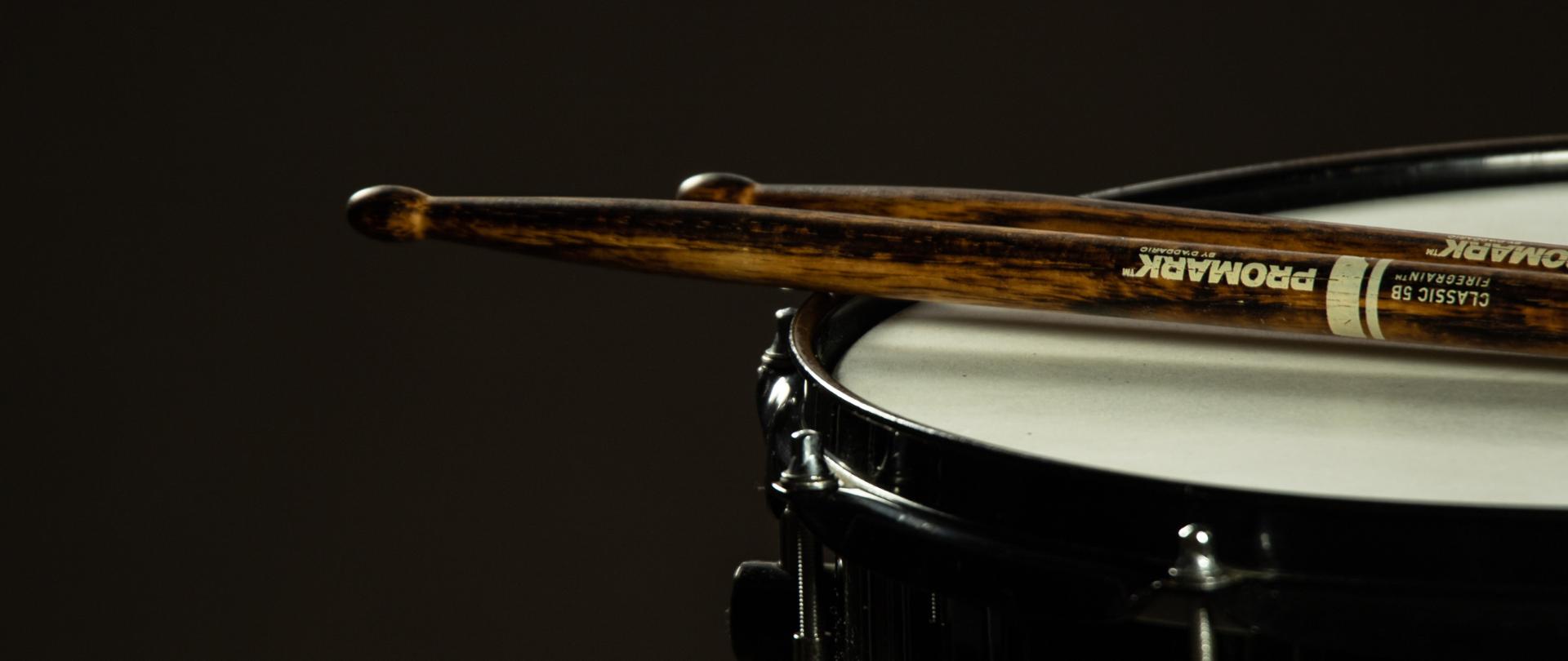 z prawej strony fragment werbla z pałeczkami perkusyjnymi, całość na ciemnym tle