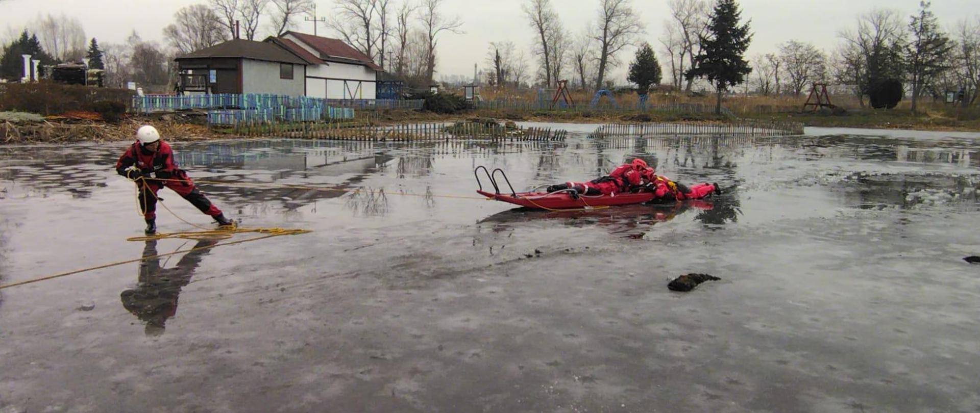 Strażacy ubrani w skafandry nurkowe podczas szkolenia na zamarzniętym akwenie wodnym, ćwiczą wykorzystanie sprzętu do ratownictwa lodowego oraz technik ratownictwa lodowego i samoratownictwa.