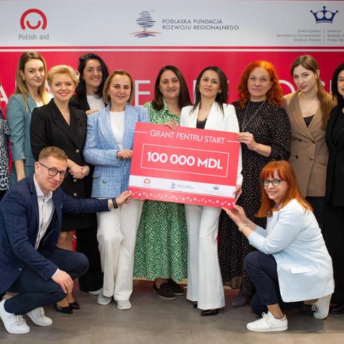 Grupa kobiet i 1 mężczyzna pozuje z dużym czekiem na kwotę 100 000 mołdawskich lei. Z tyłu na ściance widać logo Polska Pomoc, Podlaska Fundacja Rozwoju Regionalnego oraz Centrum Rozwoju i Przedsiębiorczości Polska-Mołdawia 