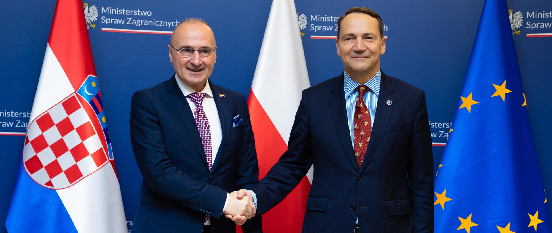Na zdjęciu jest dwóch mężczyzn w ciemnych garniturach. Podają sobie dłonie. Patrzą w kierunku obiektywu. Za nimi są trzy flagi: Chorwacji, Polski i Unii Europejskiej. 