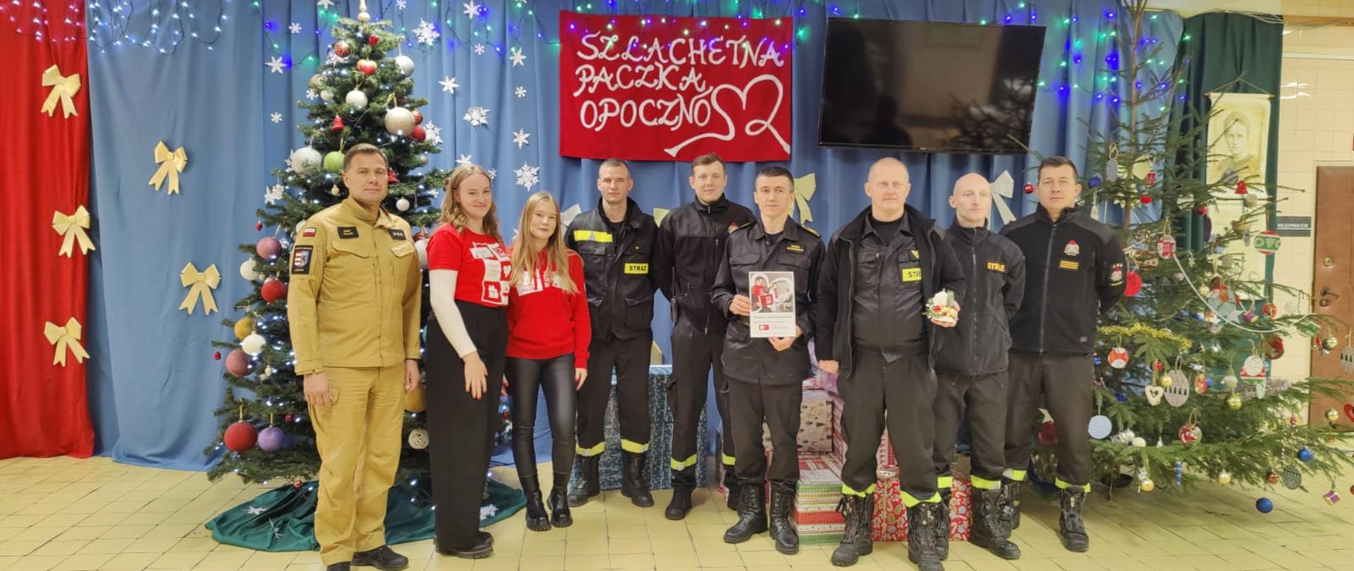 Strażacy w ubraniach koszarowych i służbowych, dwóch z nich trzyma dyplom i stroik świąteczny. Pośród nich dwie wolontariuszki szlachetnej paczki, w tle zapakowane prezenty, choinki, napis, dekoracje świąteczne na niebieskim tle. 