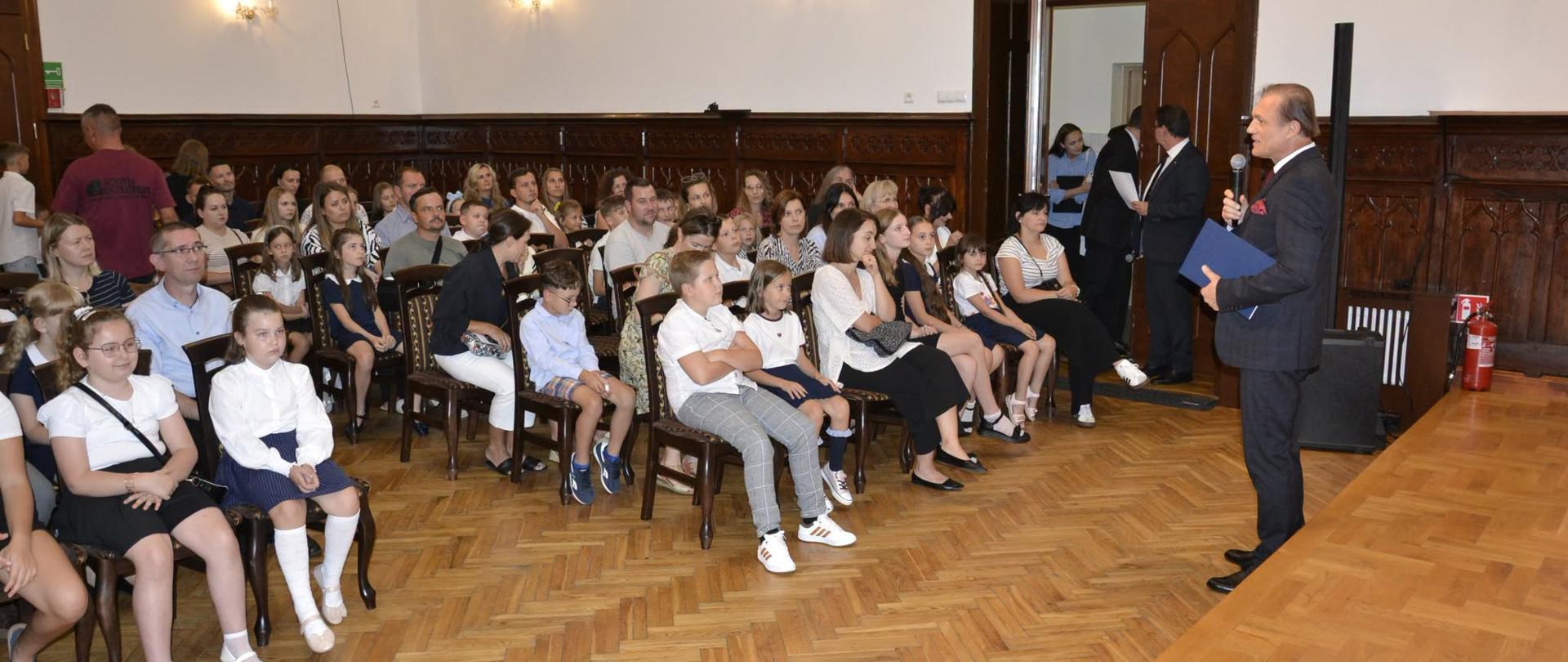 Dyrektor Ryszard Kusek przemawia z mikrofonem do uczniów klas pierwszych i ich rodziców siedzących na krzesłach w Sali Królewskiej podczas uroczystości rozpoczęcia roku szkolnego.