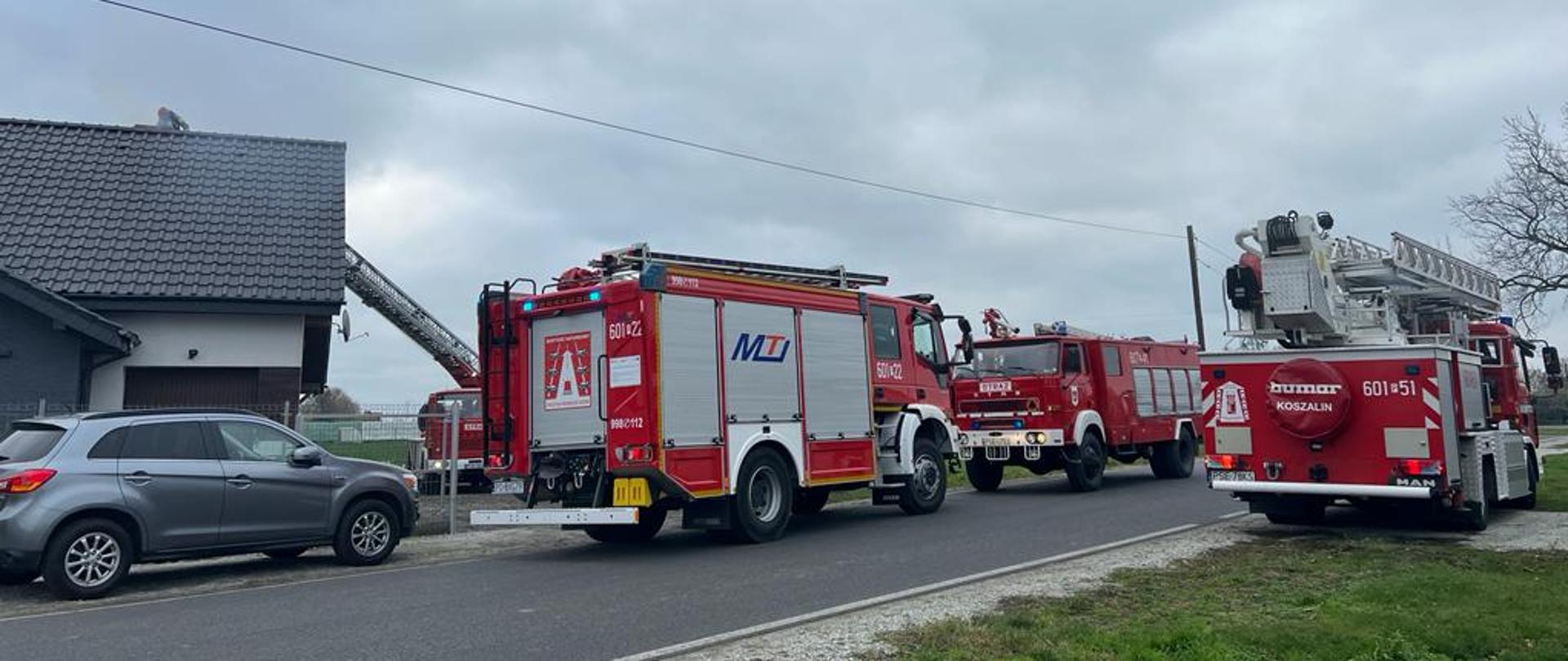Na zdjęciu 4 samochody strażackie, w tym rozstawiona drabina nad budynkiem jednorodzinnym ze strażakiem w koszu gaszącym sadze