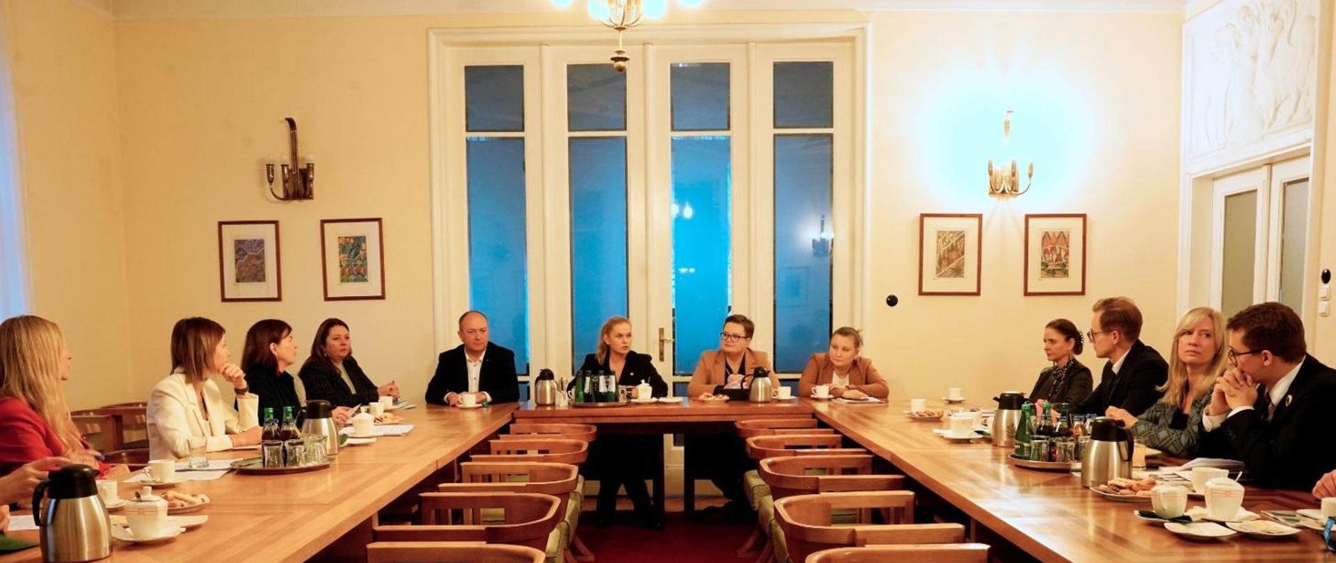 Na zdjęciu minister Barbara Nowacka rozmawia z kilkunastoma osobami, wszyscy siedzą przy stołach, z tyłu duże okno balkonowe, na ścianie wiszą obrazy.