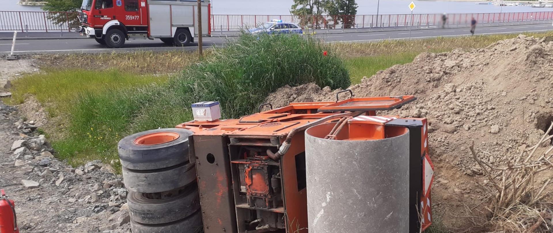 Widok walca drogowego leżącego na boku. Na drodze samochód pożarniczy.