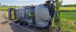 Widać uszkodzony samochód, na boku, na poboczu drogi, w pobliżu łąka