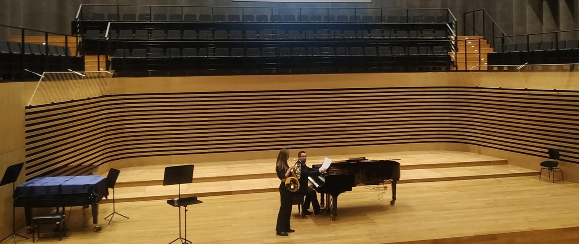 zdjęcie przedstawia waltornistkę Patrycję Kolis i akompaniatora Sławomira Kuska w czarnych strojach w dużej sali koncertowej Szkoły Muzycznej w Jastrzębiu Zdroju, sala jest utrzymana w żółtawych odcieniach drewna, a powyżej w szarych panelach wygłaszających z miejscami dla chóru.