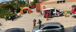 Zdjęcie przedstawia akcję ratowniczą, na której widoczni są uczestnicy ćwiczeń: strażacy, personel medyczny oraz pozoranci. Na zdjęciu widoczny jest również pomarańczowy namiot, pojazdy pożarnicze oraz samochody osobowe.