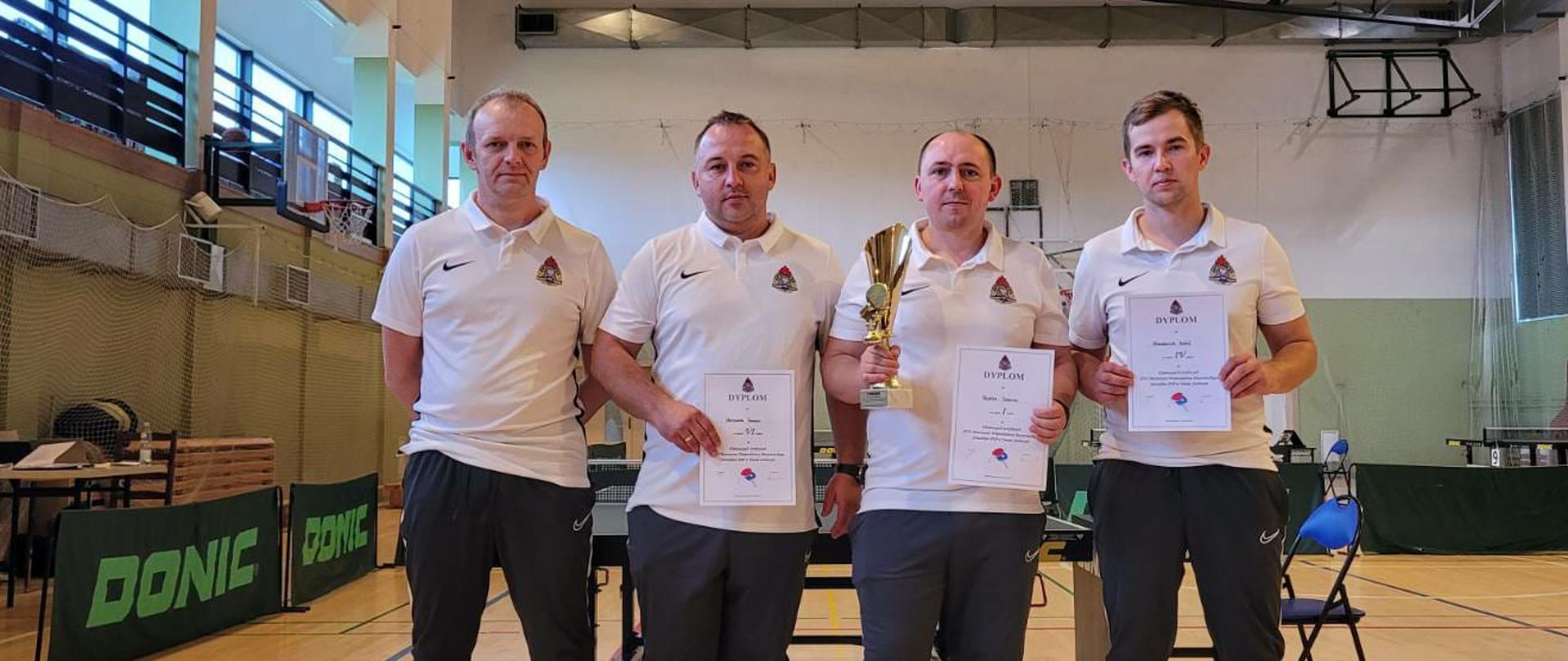 Eliminacje strefowe do XVII Mistrzostw Województwa Mazowieckiego Strażaków w Tenisie Stołowym - na zdjęciu strażacy w ubraniu dresowym stoją w szeregu na sali gimnastycznej, w rękach trzymają dyplomy.