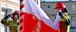 Trzech funkcjonariuszy Państwowej Straży Pożarnej uroczyście podnosi flagę państwową na maszt.