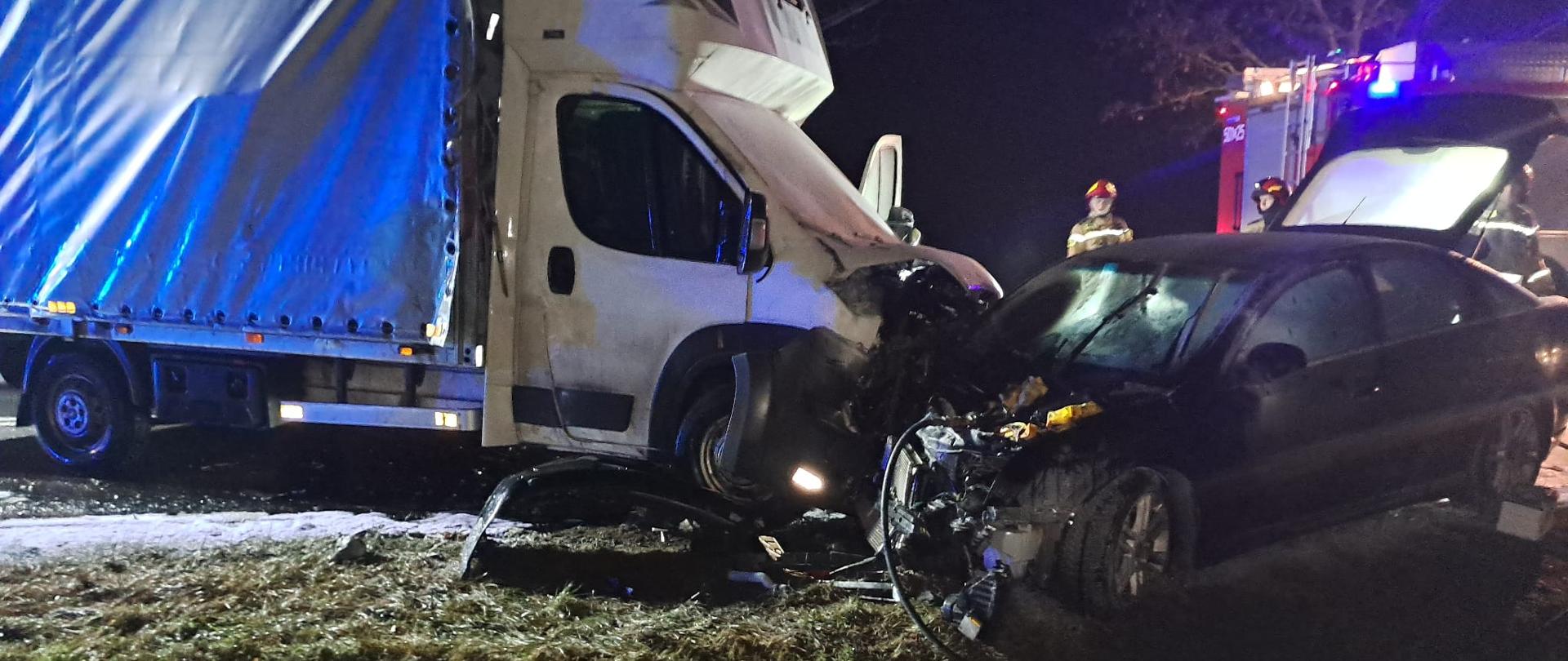 Zdjęcie przedstawia samochód dostawczy koloru białego z plandeką oraz samochód osobowy koloru czarnego po kolizji drogowej.