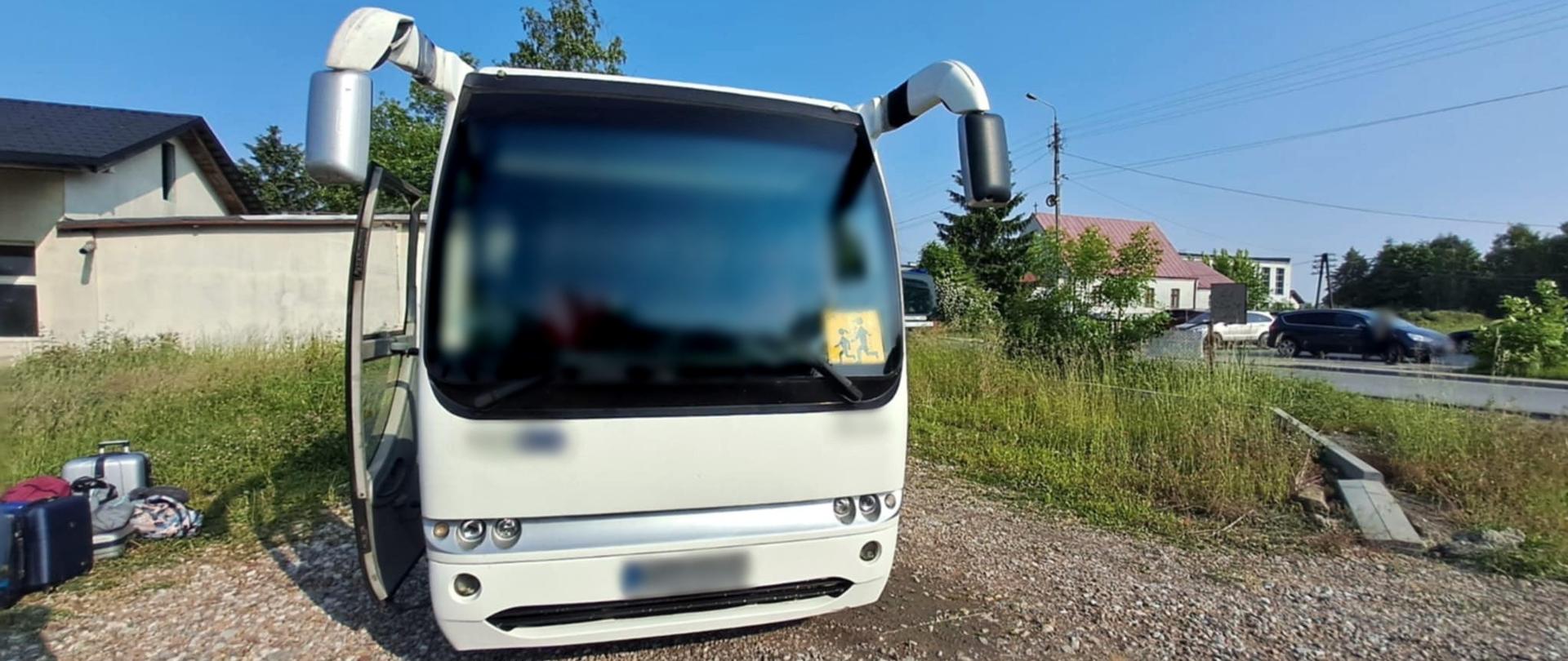 Na pierwszym planie: przód kontrolowanego autobusu turystycznego przed szkołą podstawową w Młodocinie Mniejszym koło Radomia. Obok pojazdu leżą bagaże pasażerów. W tle: oznakowany furgon mazowieckiej Inspekcji Transportu Drogowego stoi zaparkowany za kontrolowanym autokarem. 