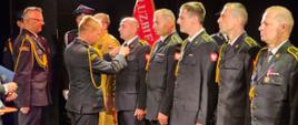 Strażacy stojący w rzędzie w galowych mundurach podczas dekoracji przez dwóch innych strażaków.
