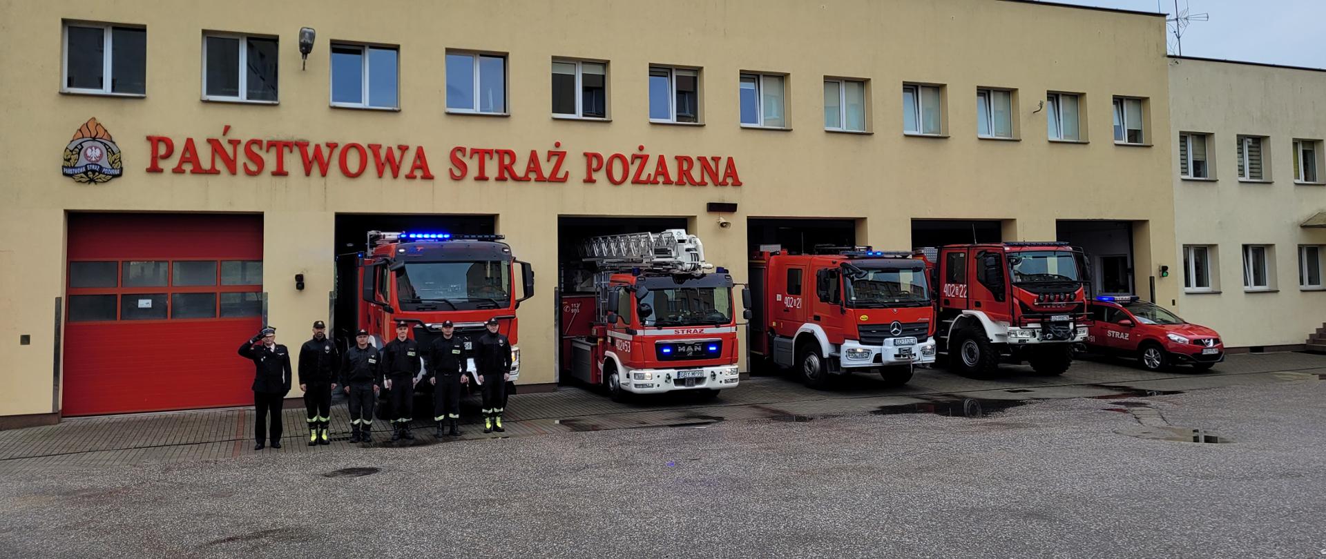 Na zdjęciu widać ustawione przed garażami samochody pożarnicze z włączonymi sygnałami świetlnymi oraz ustawionych z boku samochodów strażaków.
