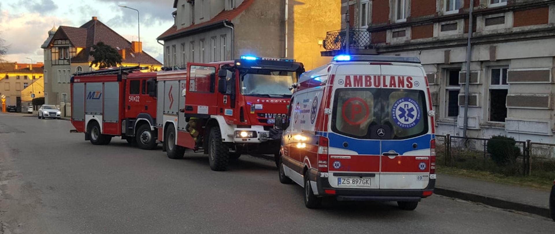 Pożar mieszkania w miejscowości Barwice - dwa samochody pożarnicze koloru czerwonego i karetka koloru białego stoją na jezdni przed kamienicą.