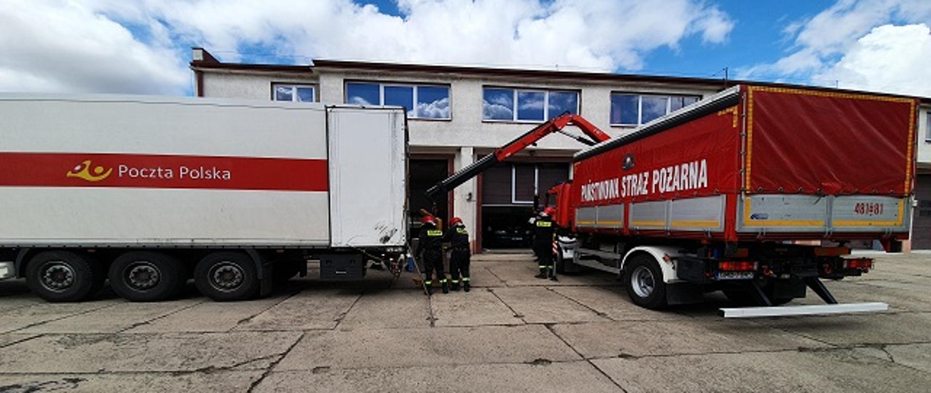 Na placu przed strażnicą stoi po lewej samochód ciężarowy dostawczy poczty polskiej, po prawej samochód dostawczy Państwowej Straży Pożarnej. Trwa wyładunek środków ochrony indywidualnej. Asystują strażacy w hełmach.
