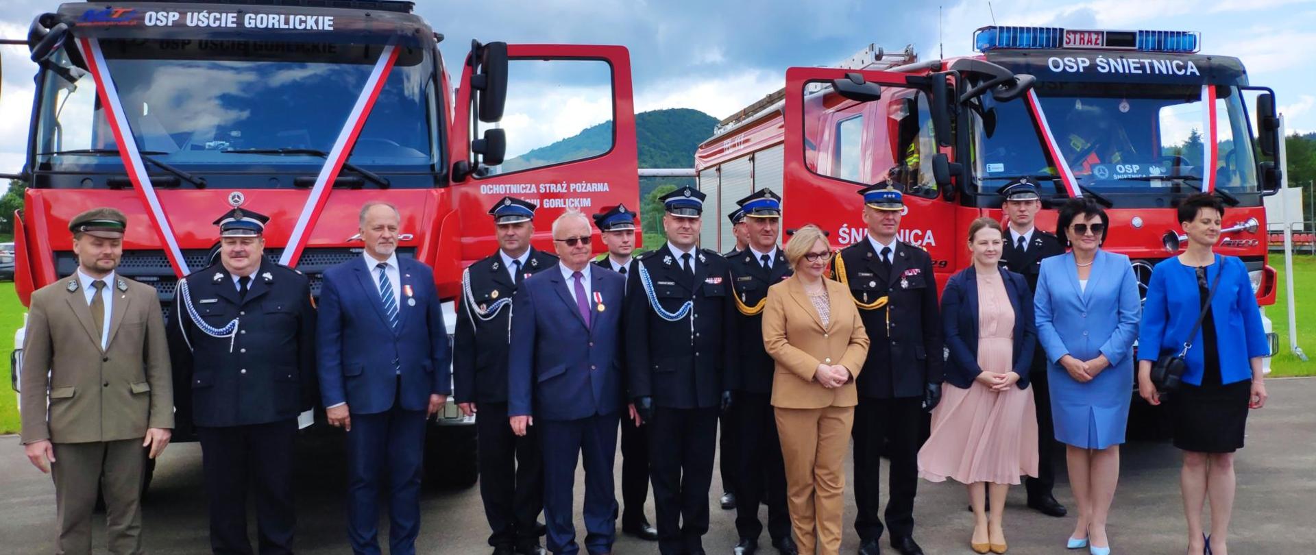 Na zdjęciu przed przekazanymi podczas uroczystości samochodami pożarniczymi koloru czerwonego stoją zaproszeni goście w tym strażacy z jednostek OSP oraz PSP w umundurowaniu galowym granatowym w czapkach rogatywkach oraz zaproszeni goście w ubraniach cywilnych. 