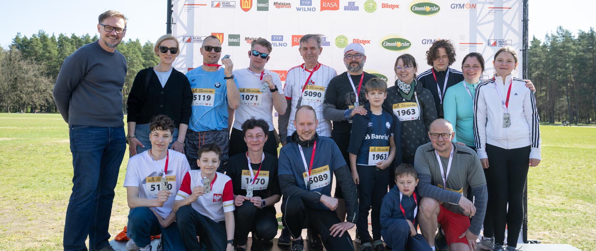 Duża grupa uczestników pozuje do wspólnego zdjęcia przed banerem biegu