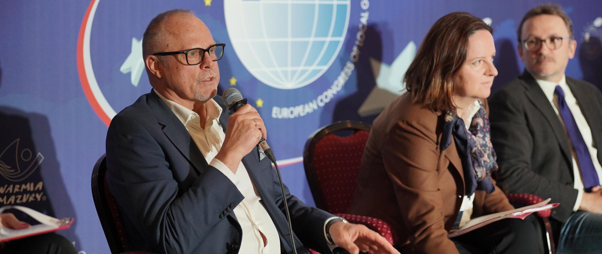 Wiceminister Jacek Protas z mikrofonem. Obok siedzą osoby.