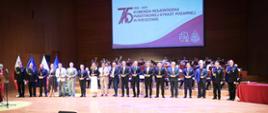 Grupa wyróżnionych osób w eleganckich strojach i mundurach stoi na scenie z nagrodami w dłoniach. W tle orkiestra i ekran jubileuszowy KW PSP w Rzeszowie.
