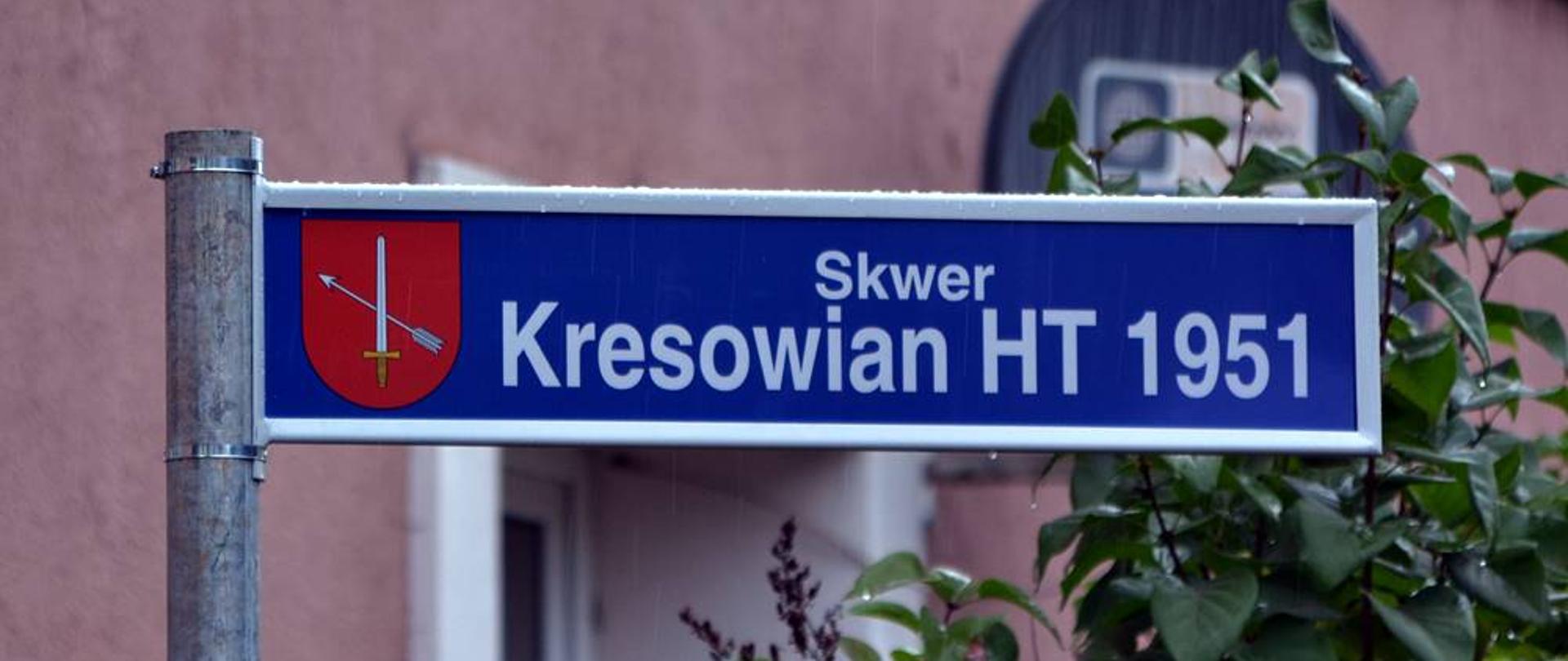 Zdjęcie przedstawia znak z napisem na niebieskim tle "Skwer Kresowian HT 1951" W tle widać rośliny oraz budynek z różową elewacją. 