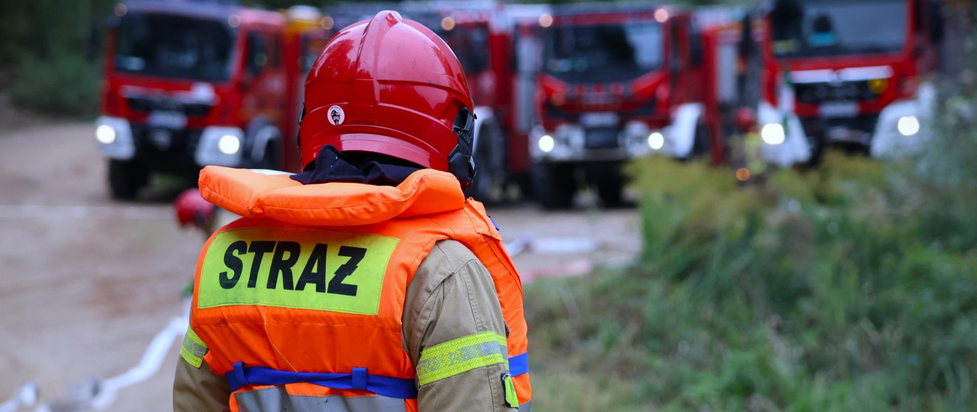 Ćwiczenia ratownicze Modułu GFFFV Białystok pod kryptonimem kryptonimem „DUŻA WODA 2025” - strażak w zabezpieczeniu, w tle samochody strażackie i linie gaśnicze 