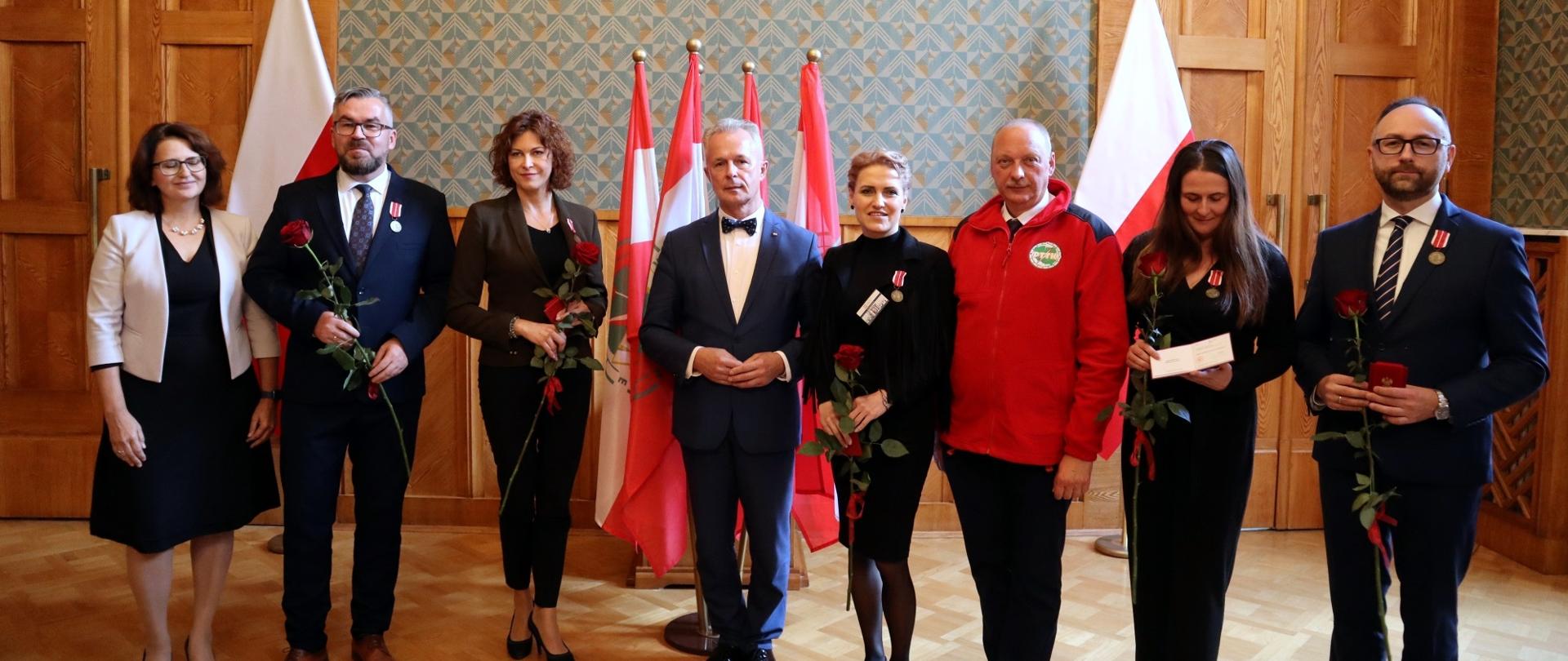 W sali z wyłożonymi drewnianymi panelami ścianami stoi w szeregu grupa ośmiu osób, w rękach trzymają po jednej czerwonej róży, za nimi pod ścianą polskie flagi.