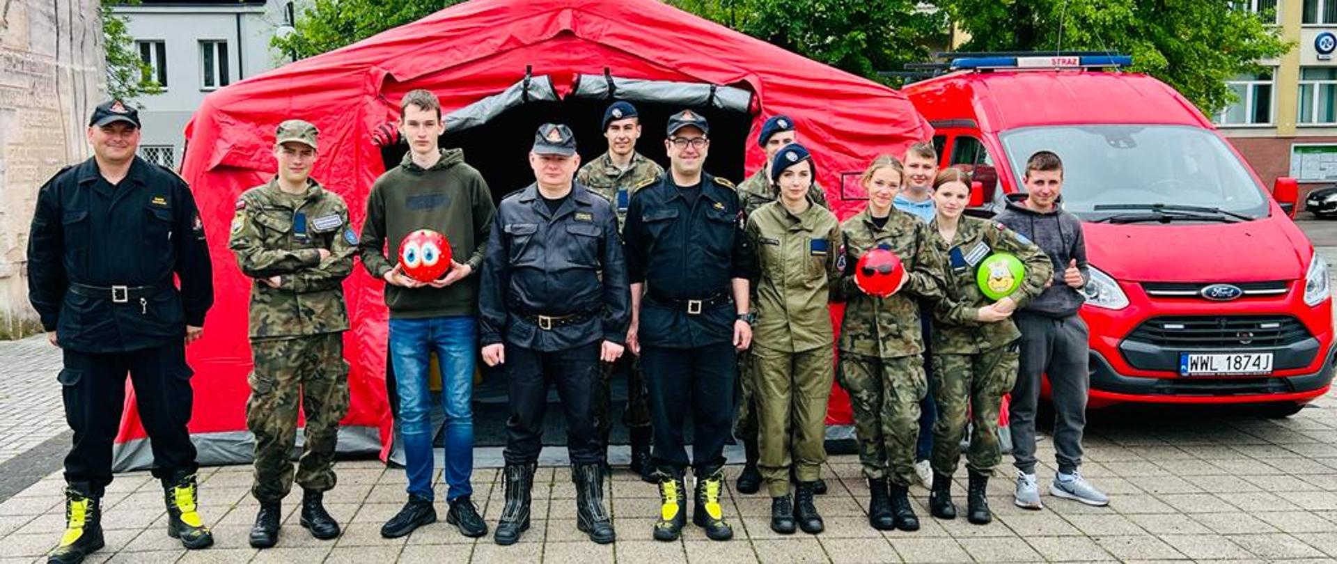 Zdjęcie przedstawia grupę młodzieży z liceum mundurowego stojących wraz ze strażakami przed czerwonym pneumatycznym namiotem strażackim. Po prawej stronie namiotu stoi czerwony bus z niebieską belką sygnałową na dachu. W tle zdjęcia widoczne są wysokie drzewa.