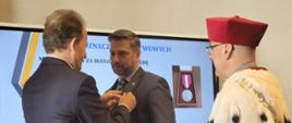 Wiceminister Bernacki przypina medal do marynarki mężczyzny w szarym garniturze, obok stoi mężczyzna w czerwonym ceremonialnym stroju z gronostajowym kołnierzem.
