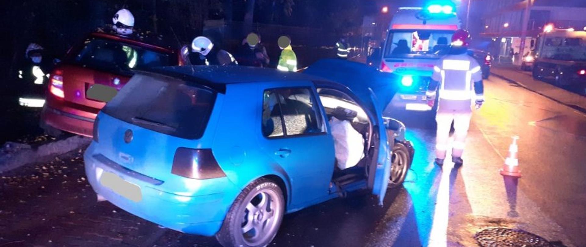Zdjęcie przedstawia wypadek dwóch samochodów osobowych. Na pierwszym planie widzimy na środku niebieski samochód osobowy, po lewej pomarańczowy samochód osobowy oraz grupę strażaków w ubraniach specjalnych i hełmach. W dalszym planie widzimy stojącą karetkę pogotowia ratunkowego. Po prawej stronie droga zabezpieczona stożkami ostrzegawczymi Zdarzenie w porze nocnej.
