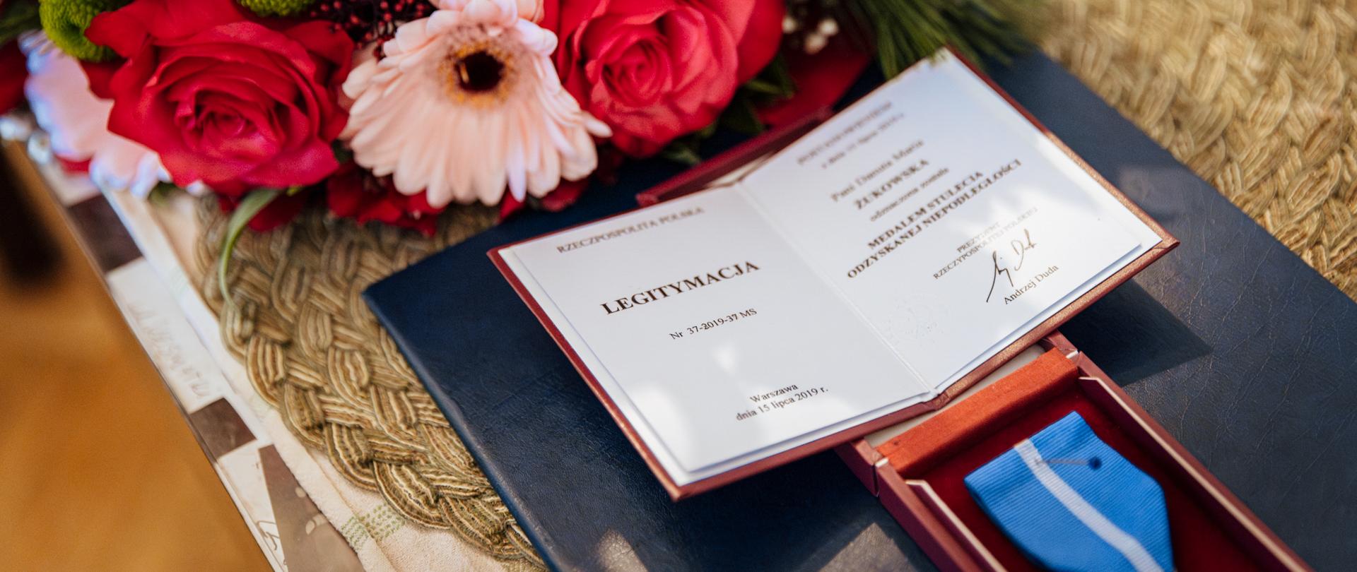 Na zdjęciu legitymacja potwierdzająca otrzymanie medalu oraz różowe kwiaty 