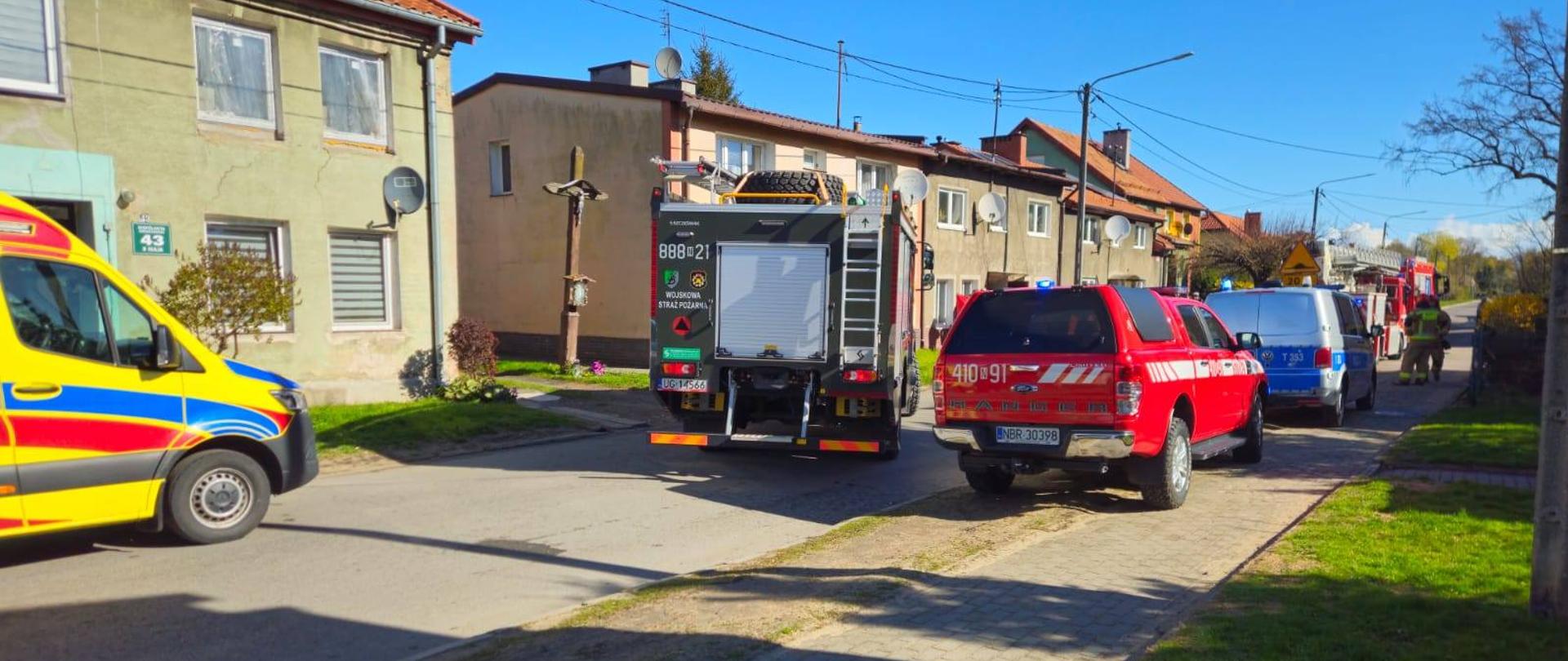 Droga osiedlowa. na drodze i poboczu ustawione pojazdy ratownicze. Z lewej strony widać karetkę pogotowia, na środku wozy strażackie i dalej radiowóz policji. Słonecznie.