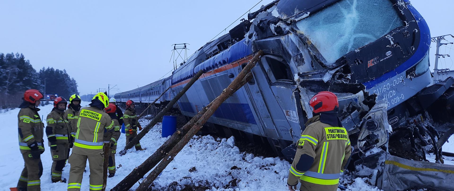 Na zdjęciu przechylona na bok lokomotywa wykolejona z boku podparta drewnianymi słupami. Wokoło strażacy PSP i OSP. Dookoła leży śnieg.
