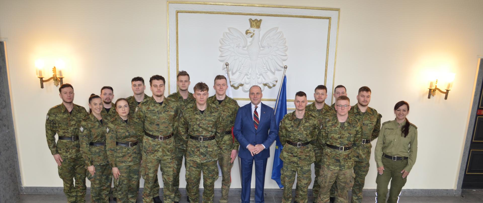 Grupa żołnierzy w mundurach wojskowych stoi w rzędzie razem z mężczyzną w garniturze pośrodku; za nimi widoczny jest biały orzeł na ścianie oraz flaga